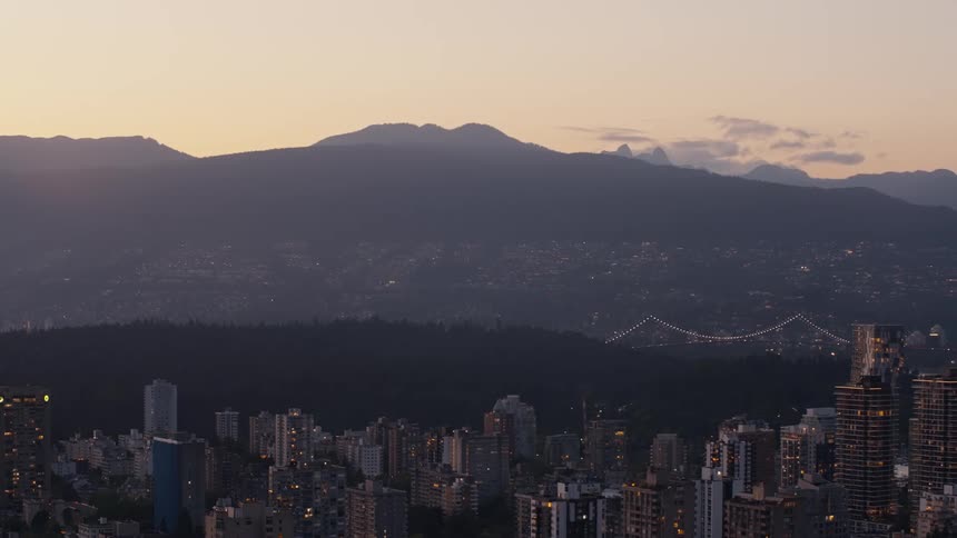 A highangle static shot captures a sprawling city skyline at dusk featuring illuminated skyscrapers in the foreground and a lit suspension bridge stretching toward a dark forest and massive mountain silhouettes under a hazy purple sky