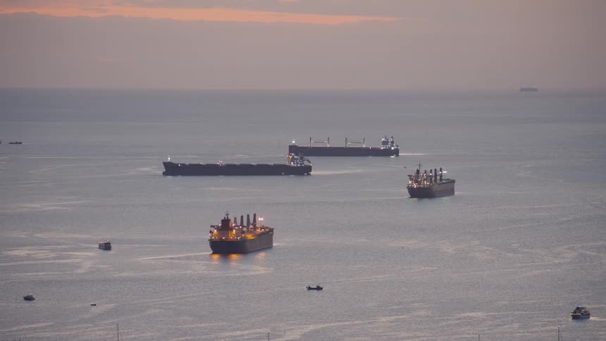 Large cargo ships and smaller vessels rest on a calm sea beneath a hazy sunset sky