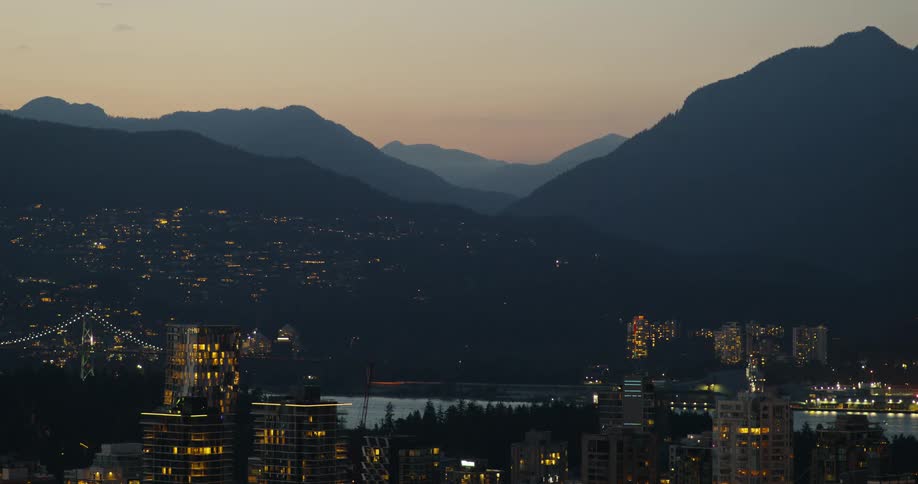 A wide view of a city skyline at dusk with glowing building windows and a suspension bridge set against dark mountain silhouettes under a dim sky