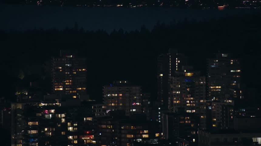 Tall buildings with apartment lights on at night against a forest backdrop silhouetted and some distant twinkle lights of buildings in the city
