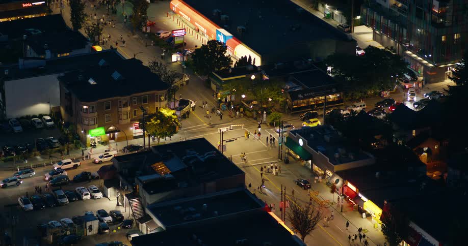 An overhead shot captures a busy city intersection at night with cars driving and pedestrians moving under streetlights and business signs