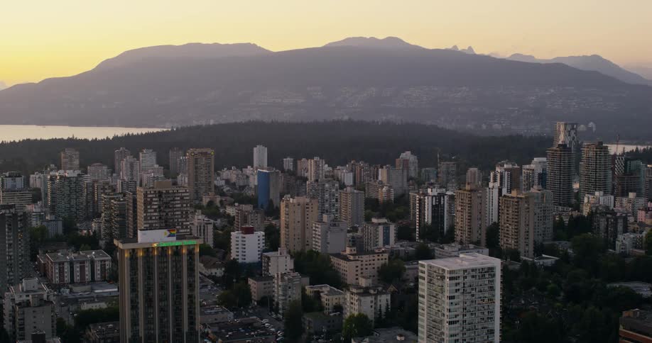 An aerial wide view captures a dense city skyline filled with tall buildings near the coast with dark mountains under a golden sunset sky