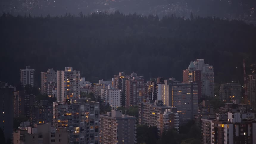 An aerial view captures a dense city district filled with tall apartment buildings with illuminated windows at night in front of a dark forested hill