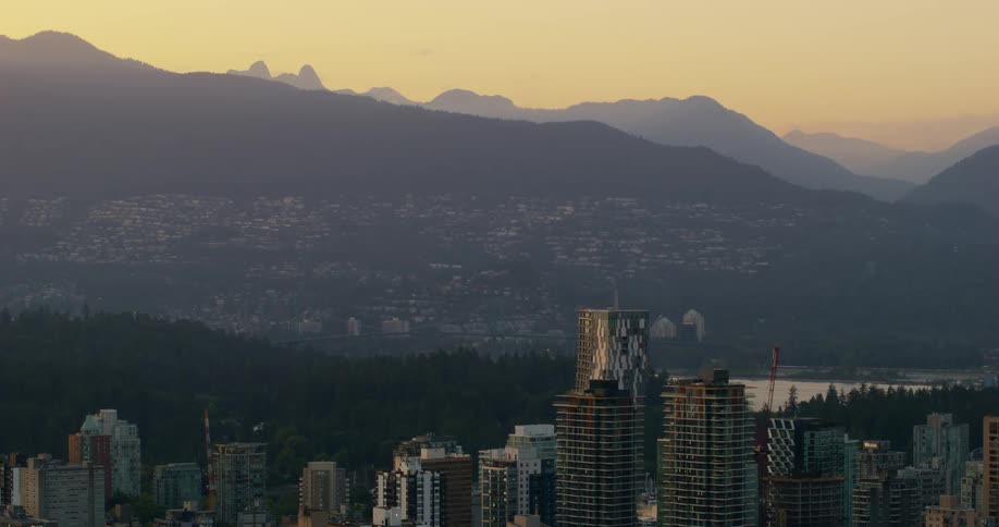 A wide view of a modern city skyline with tall buildings and a river at dusk as the sun sets behind large mountain peaks in the distance