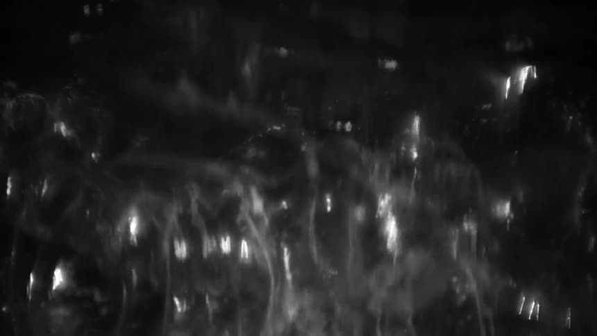 Close view of rapidly bubbling liquid in black and white with chaotic motion and bright reflections rising against a dark background