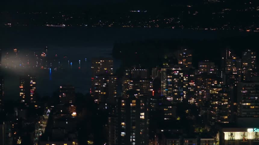 A night aerial view shows fireworks exploding over a dark bay behind a dense cluster of illuminated apartment buildings in a coastal city