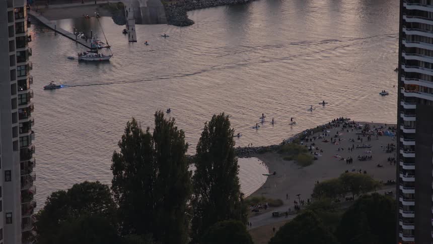 An elevated view shows a group of people on a beach next to a body of water with several boats and kayaks near the shore at dusk