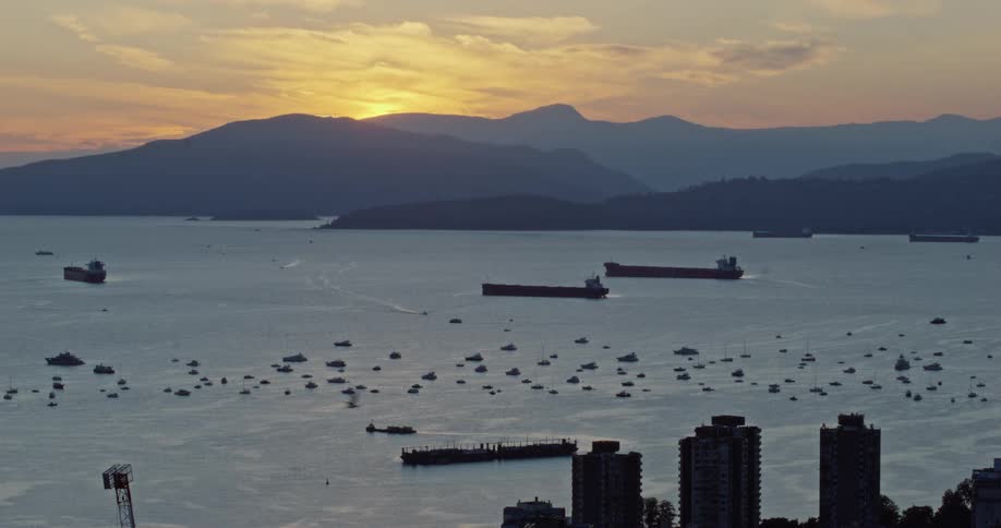 A golden sunset glows behind a mountain range overlooking a busy harbor filled with dozens of small boats and several large cargo shipsin the foreground the dark silhouettes of city highrises and a construction crane frame the coastal view