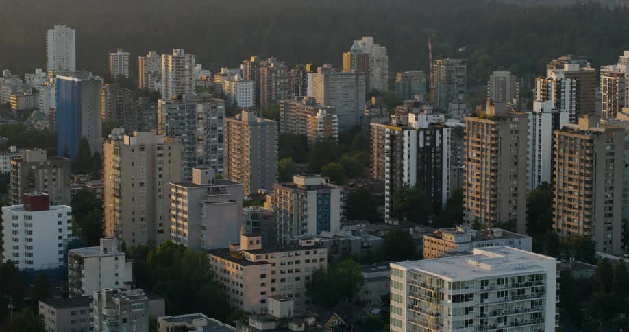 An aerial view captures a dense city district filled with tall apartment buildings glowing in golden sunset light with dark hills in the distance