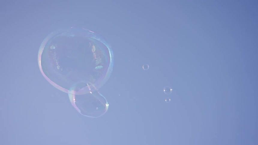 Several iridescent bubbles drift upward across a clear blue sky wobbling and distorting in the gentle breezethe sunlight catches the surface of the larger bubbles creating swirling rainbows of color before they eventually pop and disappear