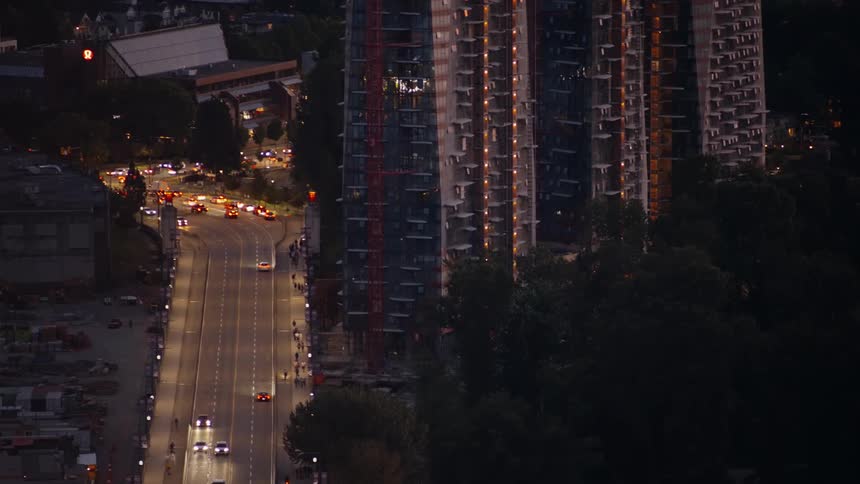 An aerial view at night captures cars driving along a lit street passing a tall modern apartment building with illuminated windows near dark trees
