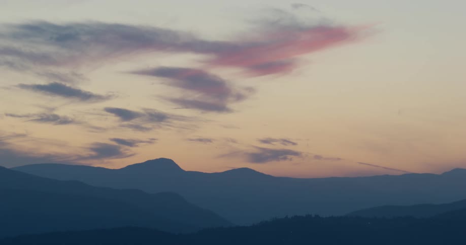Wispy pink clouds drift across a warm hazy sky above the silhouetted peaks of a serene mountain range at dusk