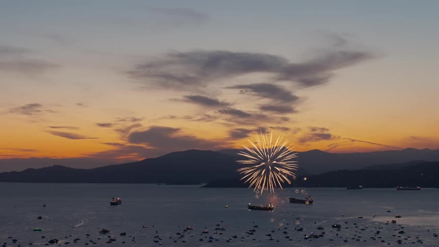 A single gold firework bursts over a calm bay dotted with cargo ships and small boats during a deep orange sunset while dark mountain silhouettes and a wispy cloud formation frame the horizon