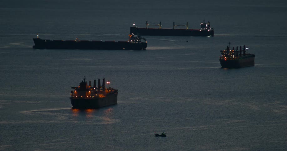 Several large dark cargo ships with glowing lights are positioned on the open water at dusk with a small boat visible in the foreground