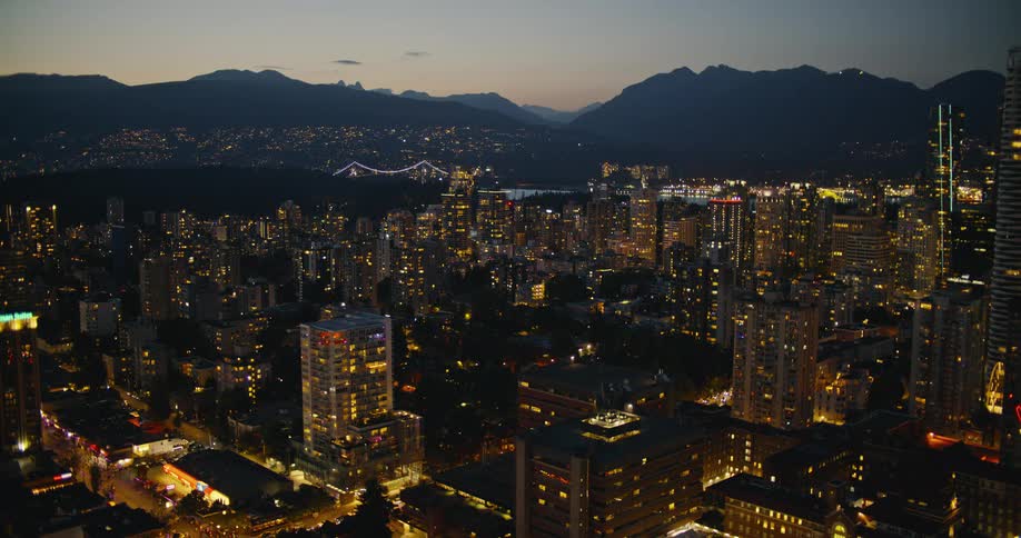 An aerial wide view captures a city skyline illuminated at night with tall buildings and a distant lit bridge in front of dark mountains under a twilight sky