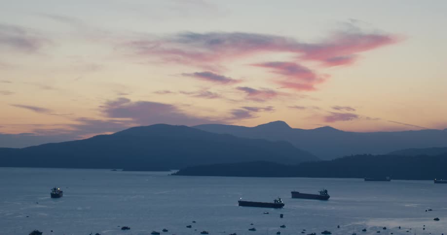 A distant view shows several large cargo ships anchored in a wide bay during a calm sunset as pink and purple clouds drift over silhouetted mountains