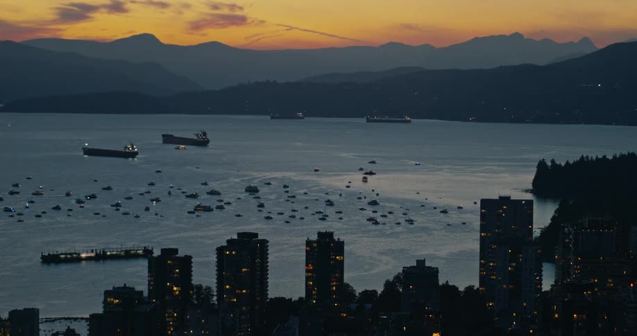 Highangle view of a coastal city at dusk showing lit highrises in the foreground while numerous boats and large cargo ships anchor in the bay against a backdrop of orange sunset skies and dark mountain ranges