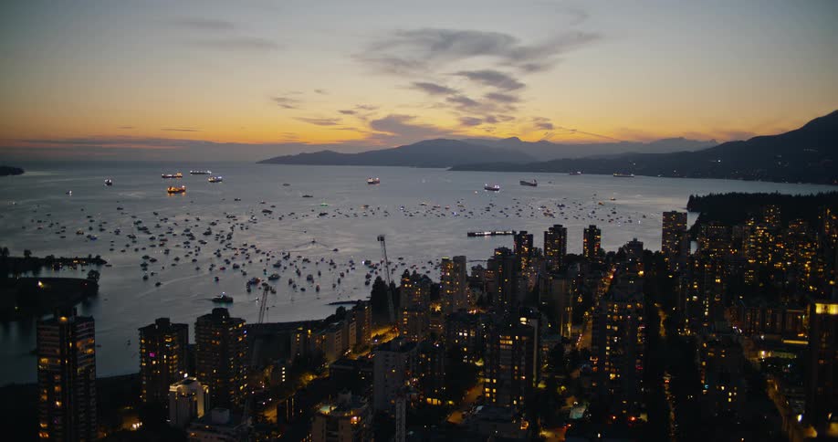 There is a large beautiful costal city beside the ocean that is full of boats and everything is lit up in the darkening sky
