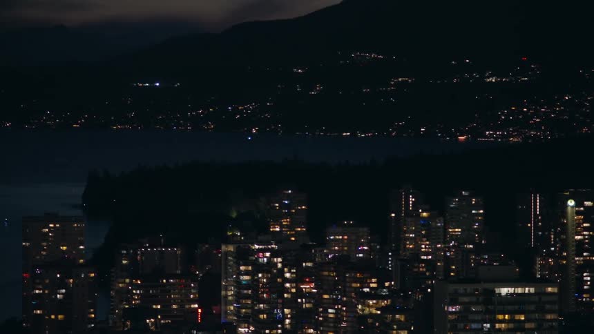 A static highangle night shot captures a dense cluster of illuminated apartment buildings and skyscrapers situated along a dark waterfront with the silhouettes of mountains and scattered lights visible in the distant background across the bay