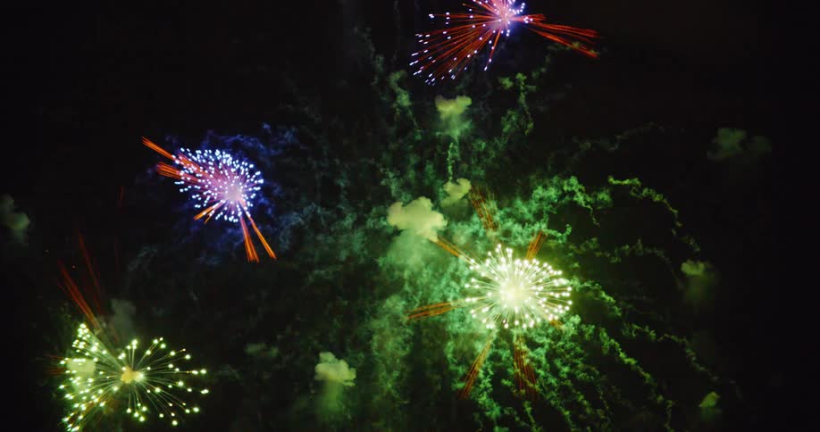 Bright colorful fireworks explode against a dark night sky illuminating the clouds with red and gold light