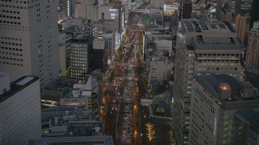 An aerial view captures a busy city avenue at night filled with cars and lined with illuminated trees between tall modern buildings