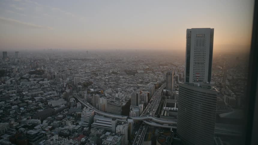 From a highaltitude window the camera captures a wide view of a city skyline at sunset showing dense residential blocks a winding highway with moving cars and a prominent skyscraper standing against the hazy orange horizon