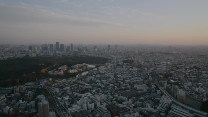 An aerial view pans across a sprawling metropolitan landscape at dusk revealing a massive dark green park nestled among a sea of lowrise buildings and distant skyscrapers under a hazy sunset sky