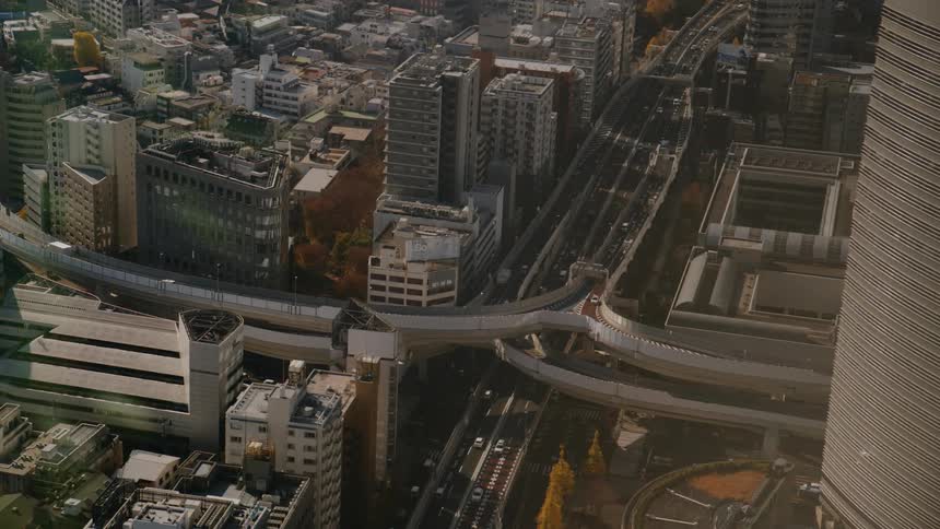 An aerial view looks down on a winding highway interchange situated between tall modern buildings with cars driving on the roads below