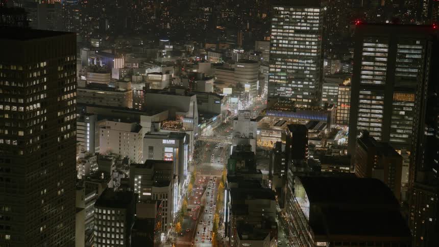 A highangle nighttime view shows a dense urban cityscape with bright street lights illuminated office buildings and a long line of cars moving slowly through a main thoroughfare lined with seasonal trees