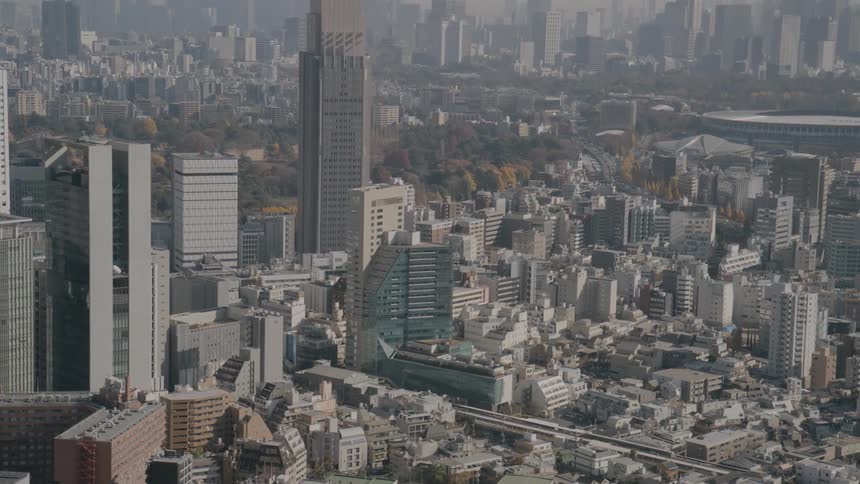 An aerial shot captures a vast hazy cityscape featuring the prominent ntt docomo yoyogi building towering over the surrounding urban density the camera pans and tilts down to reveal a complex network of tightly packed residential and commercial building