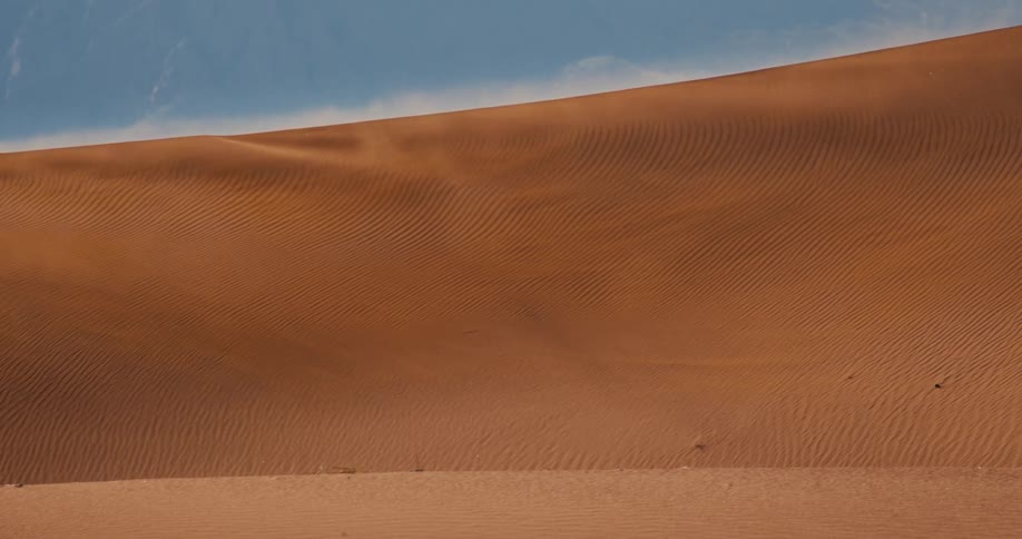 Wind blows across the sharp crest of a large orange sand dune lifting fine particles into the air while sunlight highlights the rhythmic ripple patterns covering the desert slope against a pale blue sky