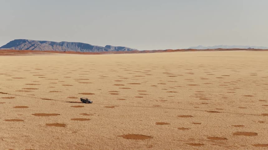 An aerial shot captures a dark offroad vehicle across a vast desert dotted with natural fairy circles toward distant blue mountains under a clear sky