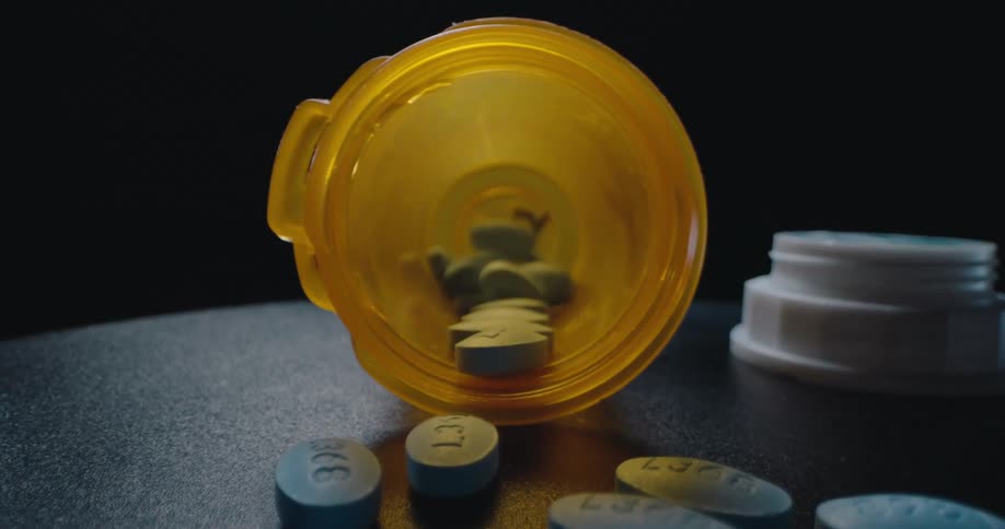 A close view of blue pills scattered on a dark surface with an open orange prescription bottle as the camera moves inside the container against a black background