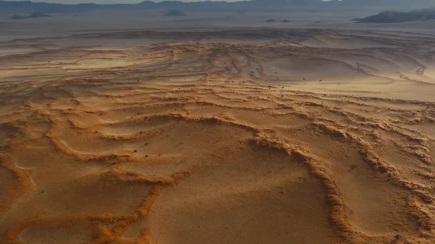 An aerial camera flies over an expansive field of orange sand dunes that create curving patterns across a dry landscape leading toward hazy mountains on the distant horizon under a soft morning light