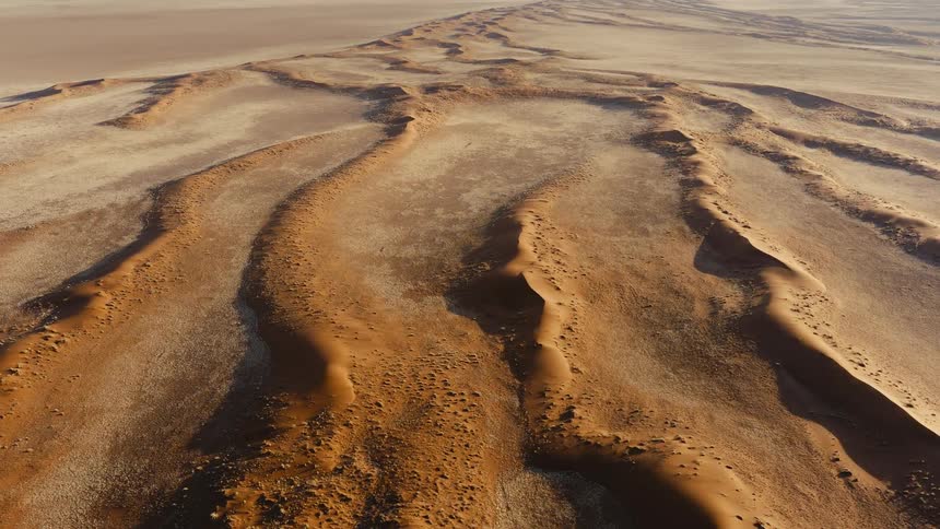 An expansive aerial view soars over a vast sundrenched desert revealing the intricate winding patterns of massive sand dunes that stretch endlessly toward the horizon