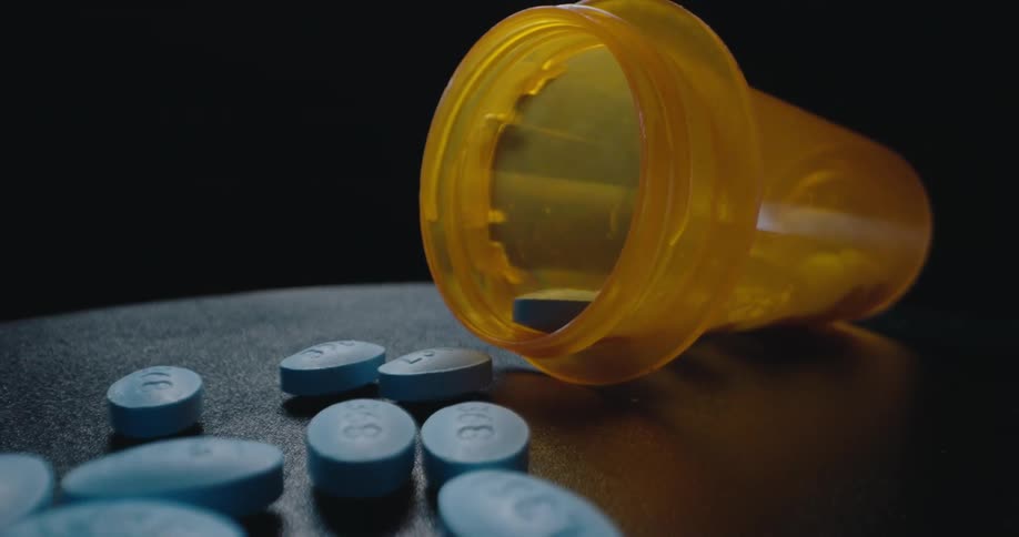 A closeup view of an orange pill bottle tipped on its side with several small blue ovalshaped pills scattered on a dark textured surface next to it