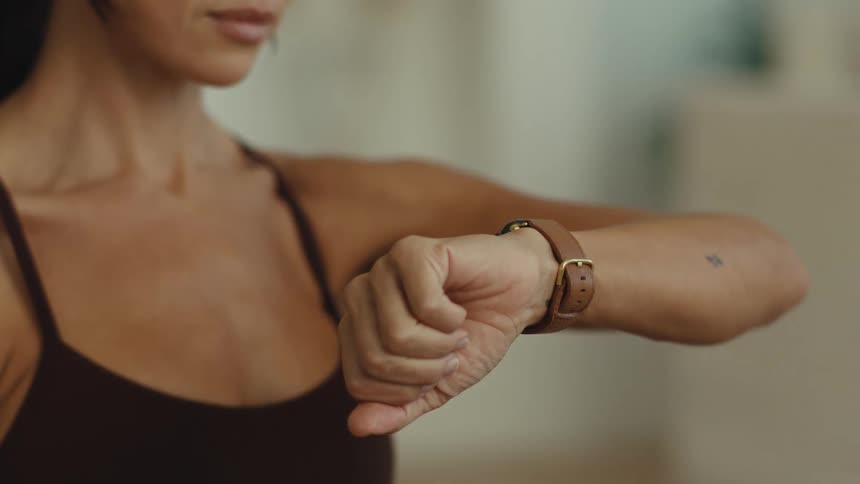 A woman wearing a brown sports bra lifts her arm to check a wristwatch with a leather strap in an indoor setting