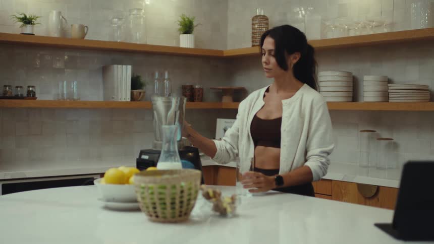 A woman wearing athletic clothing pours a green smoothie from a blender into a glass in a bright modern kitchen with wooden shelves and white counters