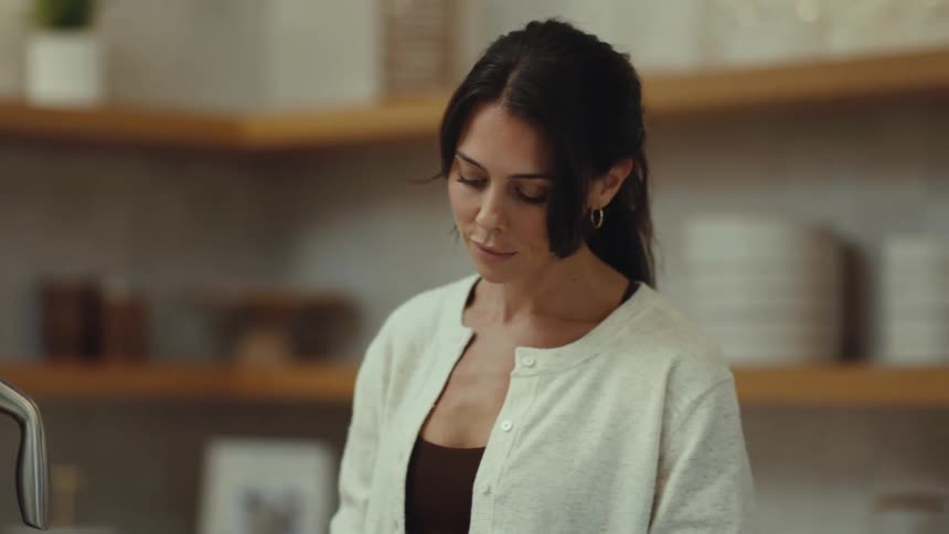 A woman with dark hair tied back stands in a kitchen looking down at a counter while she prepares food and occasionally glances toward a metal pitcher