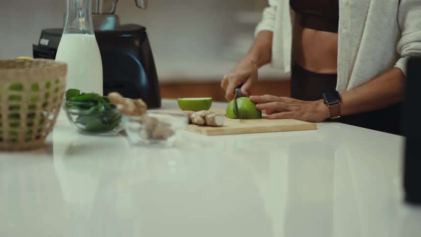 Young adult woman slicing apples and putting on a blender over a counter on a kitchen indoors