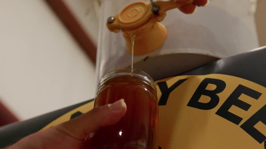 A person holds a glass jar under a yellow plastic gate valve as a stream of thick golden honey pours steadily from a large white bucket into the container