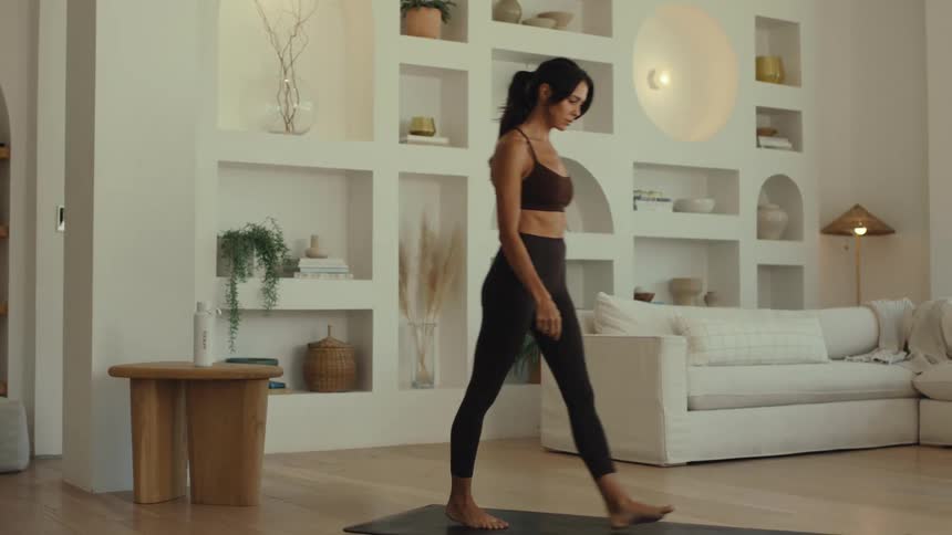 The woman walks across the room lays her yoga mat out and sits down on itshe is in a white living room with a white couch