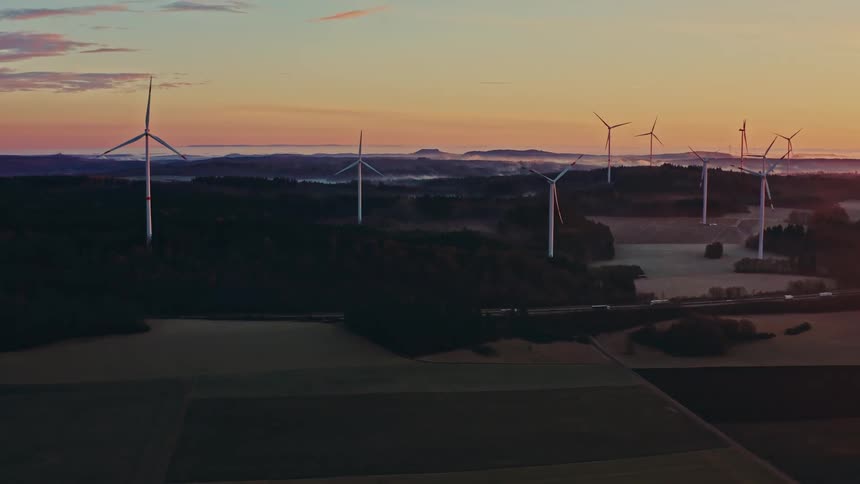 A wide aerial shot pulls back over a wind farm at sunrise its turbines spinning gently above a landscape of dark fields and forests