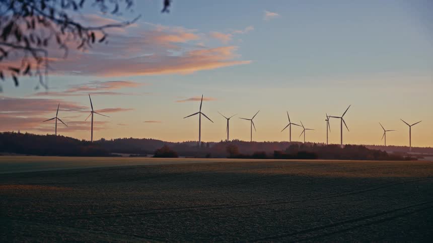 A line of wind turbines rotates slowly on a dark ridge overlooking a wide green field during a colorful sunset while silhouetted tree branches frame the top left corner of the rural landscape as the sky transitions from orange to blue