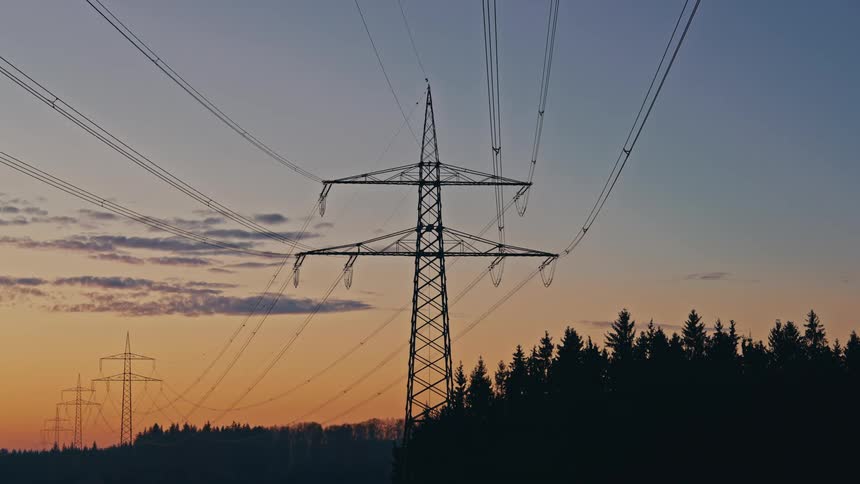 A series of highvoltage transmission towers and power lines stand silhouetted against a gradient sunset sky over a dark pine forest during the evening twilight