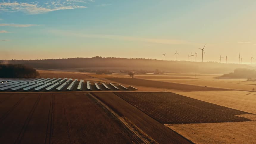 Solar panels and wind turbines operate in rural field under golden light