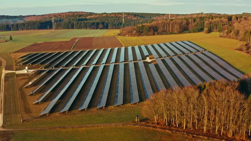 An aerial shot captures a large solar panel farm situated in a hilly rural landscape at sunset