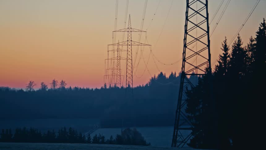 Electric transmission towers aligned over a rural landscape with silhouetted trees at dusk