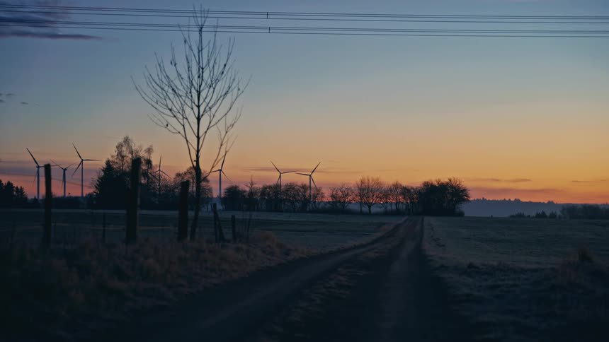 A dirt road winds through the countryside toward wind turbines at sunset