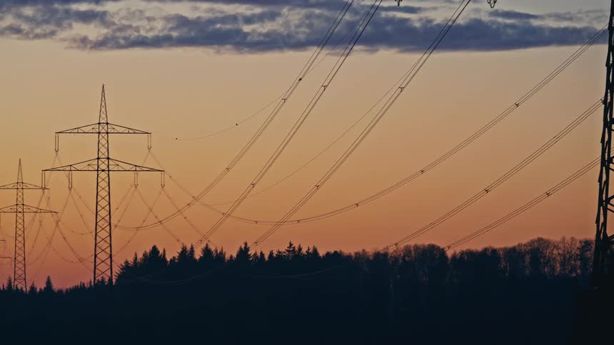 A metal power tower stands in a field next to a dark forest under a blue and orange sky at dawn